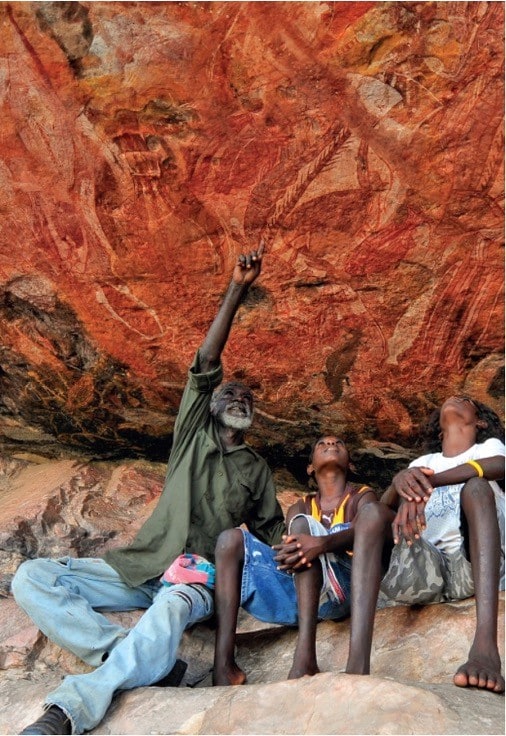 An image of an aboriginal man pointing at a rock with 2 young indigenous people
