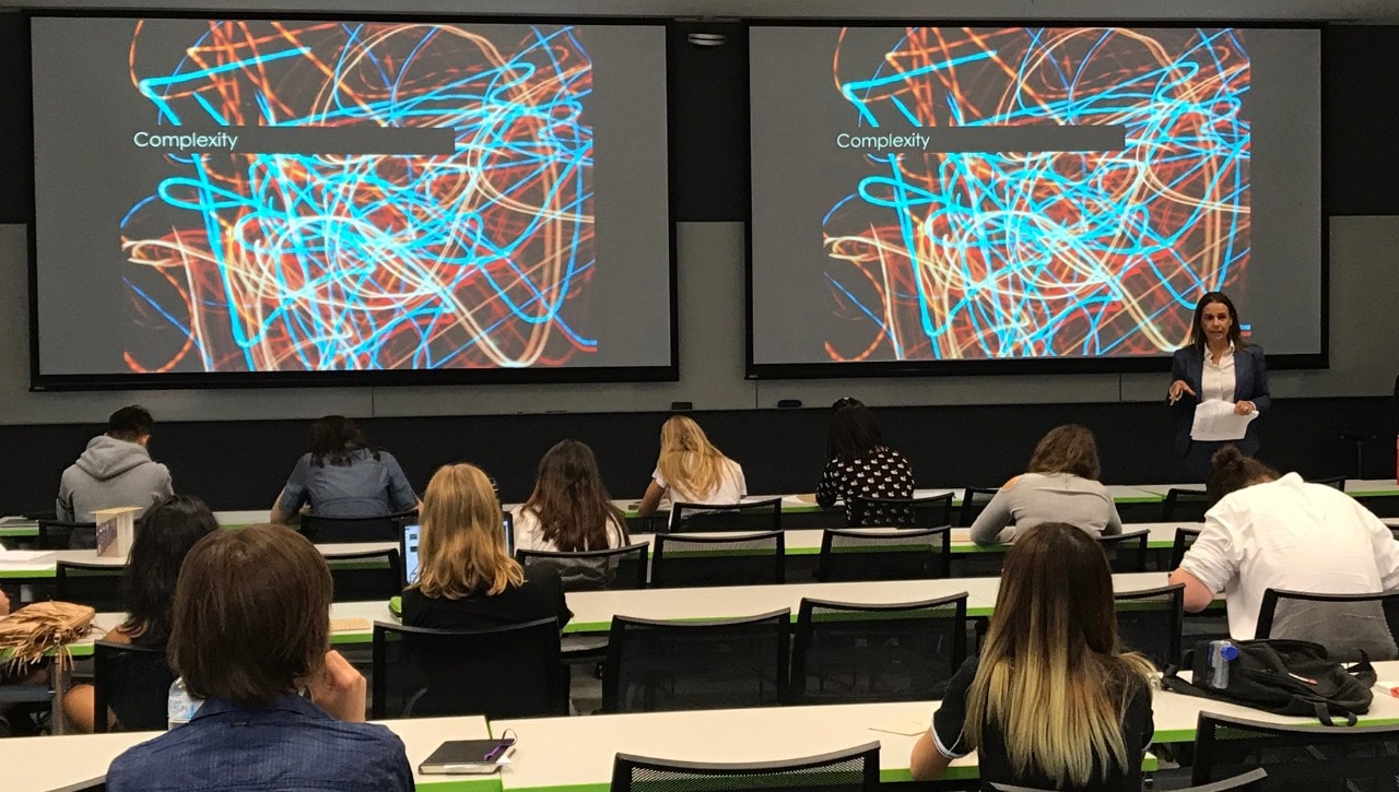 Lecture theatre with two screens displaying "complexity", female speaker, students seated, viewed from the back of the room