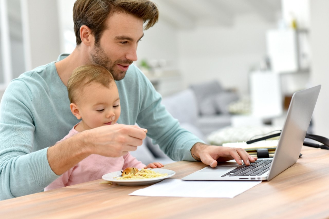 Father feeding child on lap, working on laptop