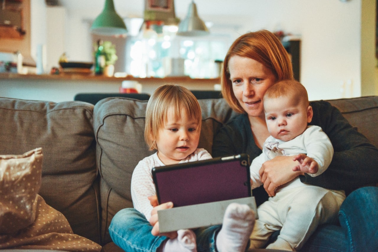 Stock image of mother on couch reading off an iPad to a toddler and baby. Photo by Alexander Dummer on Unsplash.