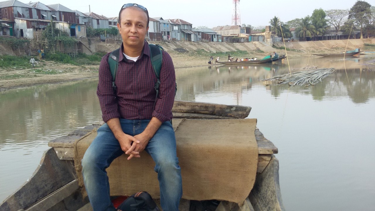 Shaymal Chowdhury sits in the back of a large, wooden boat. He is wearing jeans, a maroon jumper and a back pack, and on a river with light brown water under a pale sky. In the background are rundown buildings made of metal sheeting.