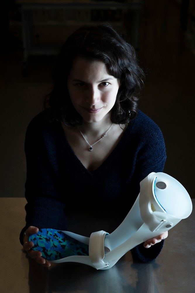 Dr Tegan Cheng sitting at a table and holding a familiar-style foot and leg brace, with straps at the ankle and knee. She is against a dark background and gently side lit. She is wearing a dark v neck top and a necklace with a small pendant.