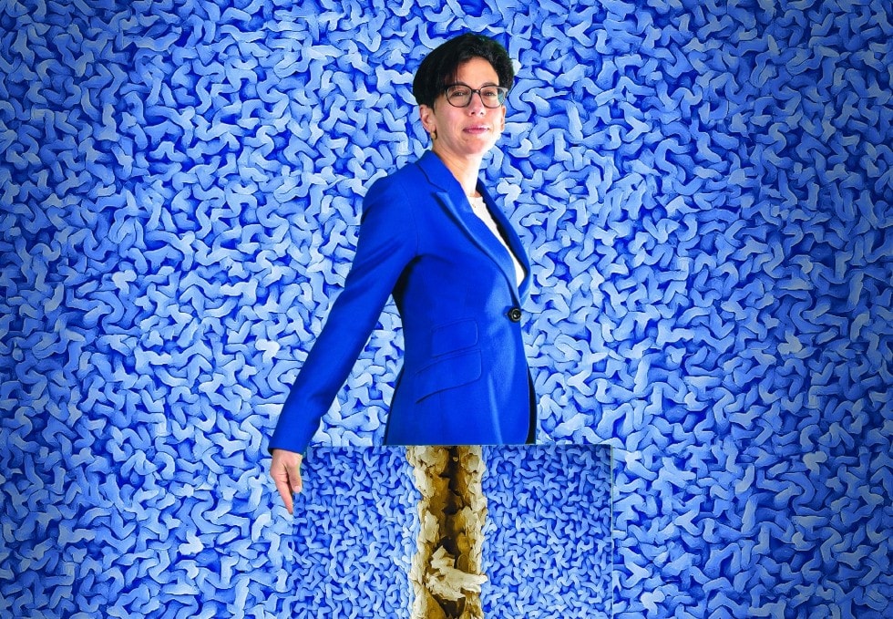 Associate Professor Chiara Neto, with large, black-rimmed glasses and a vivid blue jacket stands in front of of a gorgeously textured purple-blue wall. She is holding in front of her, a picture of the same texture but with a thick, rough brown gouge in it.