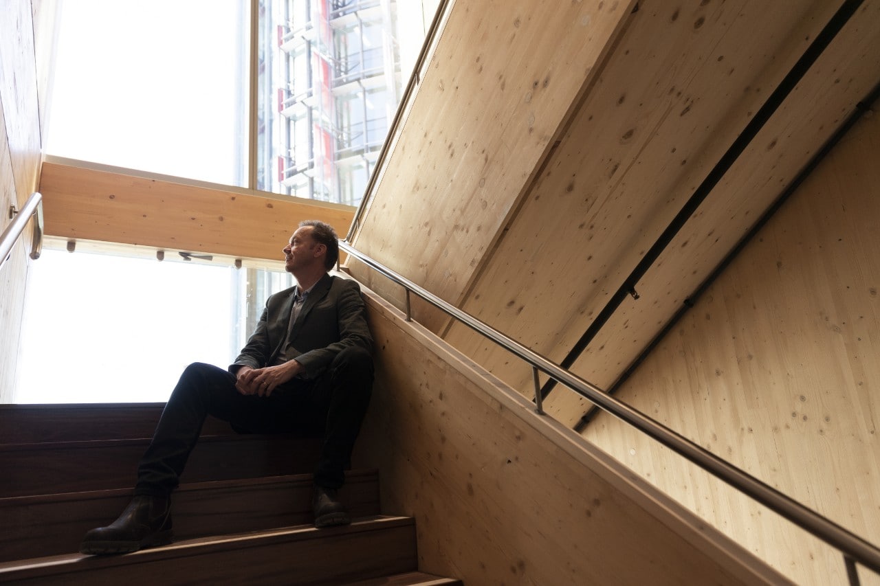 Jonathan Evens sitting in the stairwell of Daramu House. He is looking up the stairway to a window and the soft light coming in bathes the all-wood surfaces showing them as a dark beige. A neighbouring skyscraper can be seen out the window.
