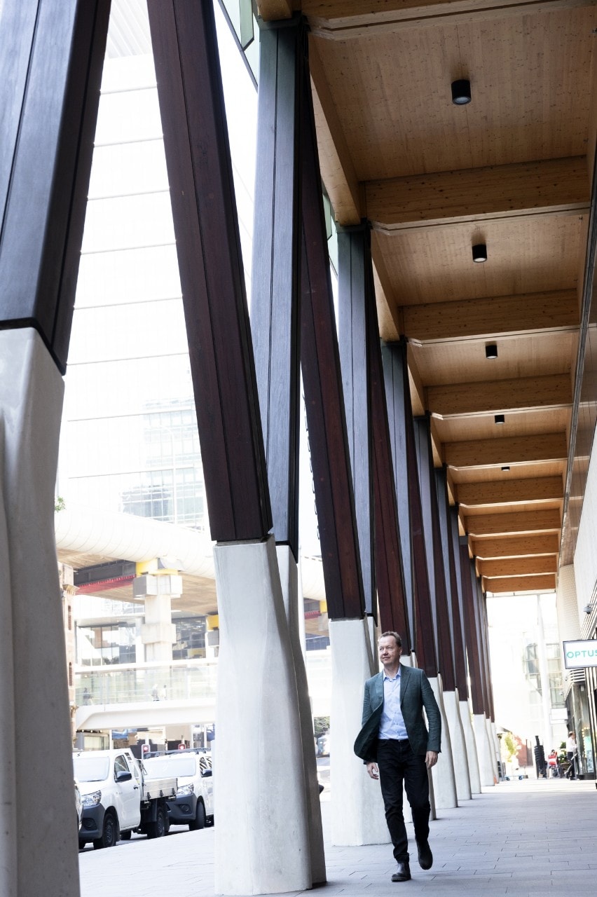 A long shot of Evans wearing a suit as he walks along the colonnade outside Daramu House. To his left we see a row of grey, concrete struts supporting the building, which feed into dark brown, wooden Vs which connect with the colonnade roof. The roof is also wood but paler in colour.