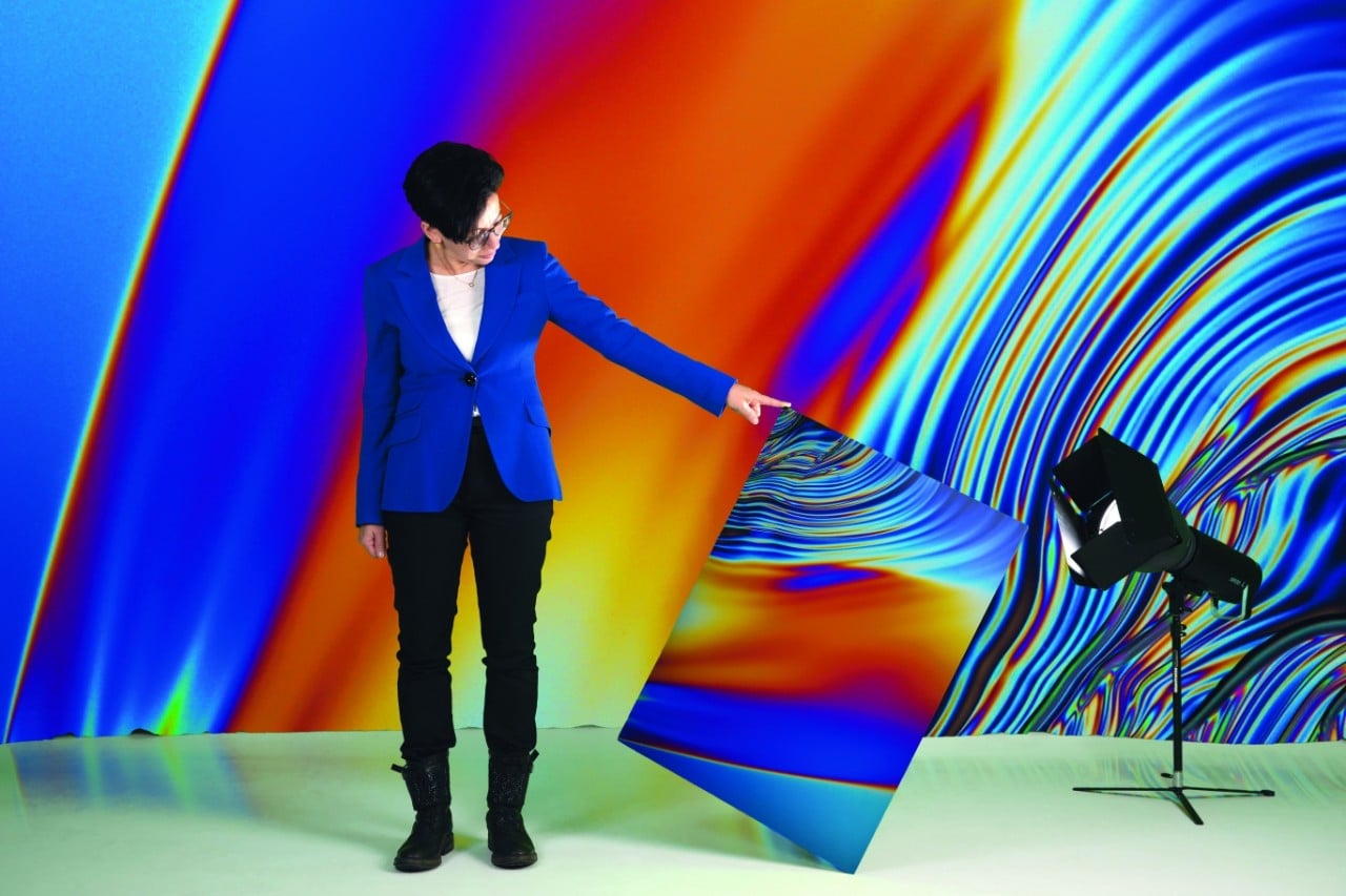 Chiara Neto in a swirl of rainbow colours on the wall behind her and on a large sheet she is holding. A photographic light is on the floor to her right.