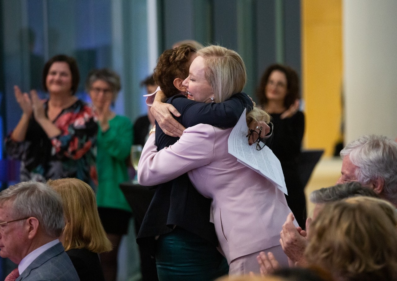 Charlotte Wood and Judy Harris standing and embracing each other in a hug in a room full of people who are clapping.