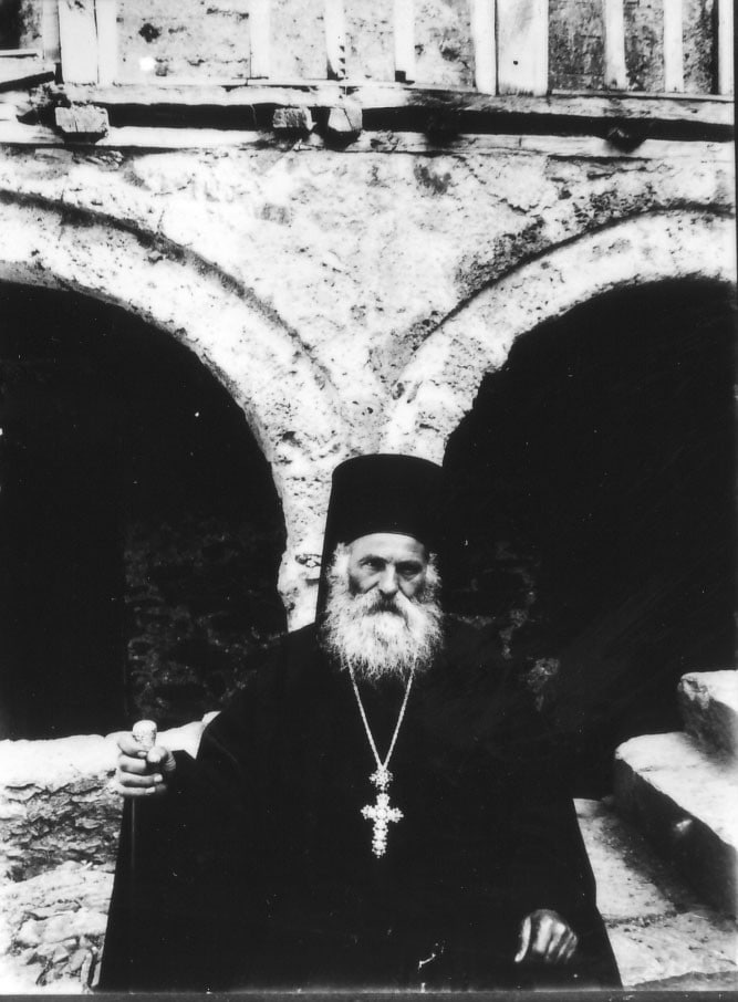 Black and white photo of a monk in front of a monastery in Pheneos, Greece. He is dressed in black and has a long beard, wears a crucifex.