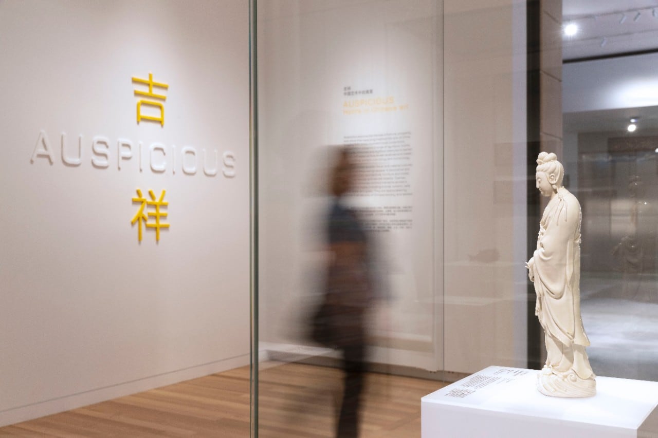 A visitor to the gallery looks at the white sculpture of Guanyin standing on a wave. Behind them is the name of the exhibiton: Auspicious. 