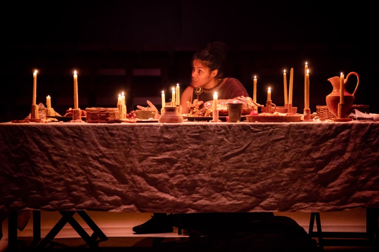 photo of actor Zahra Newman sitting at a table on stage covered in candles for dinner