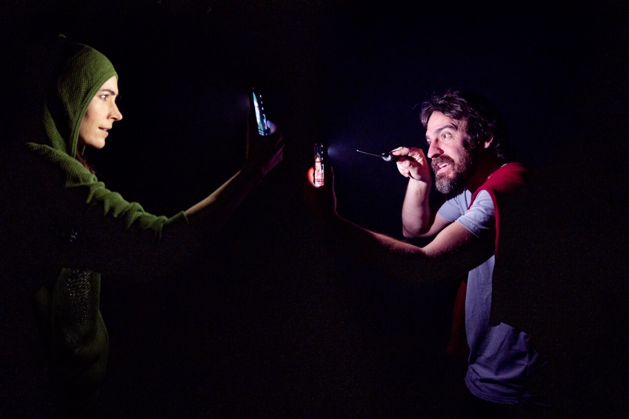 photo of two actors staring at their mobile phones in almost total darkness on stage. One is Caska and the other is Cassius.