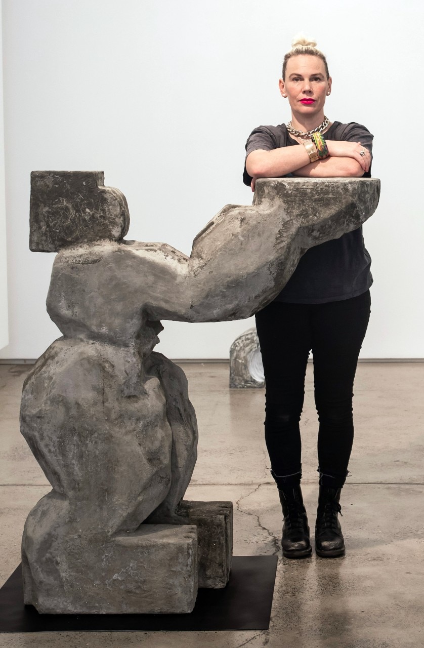 Sanne standing with one of her sculptures, it is made of stone and is as tall as her, it looks like an abstract female figure holding up a bowl