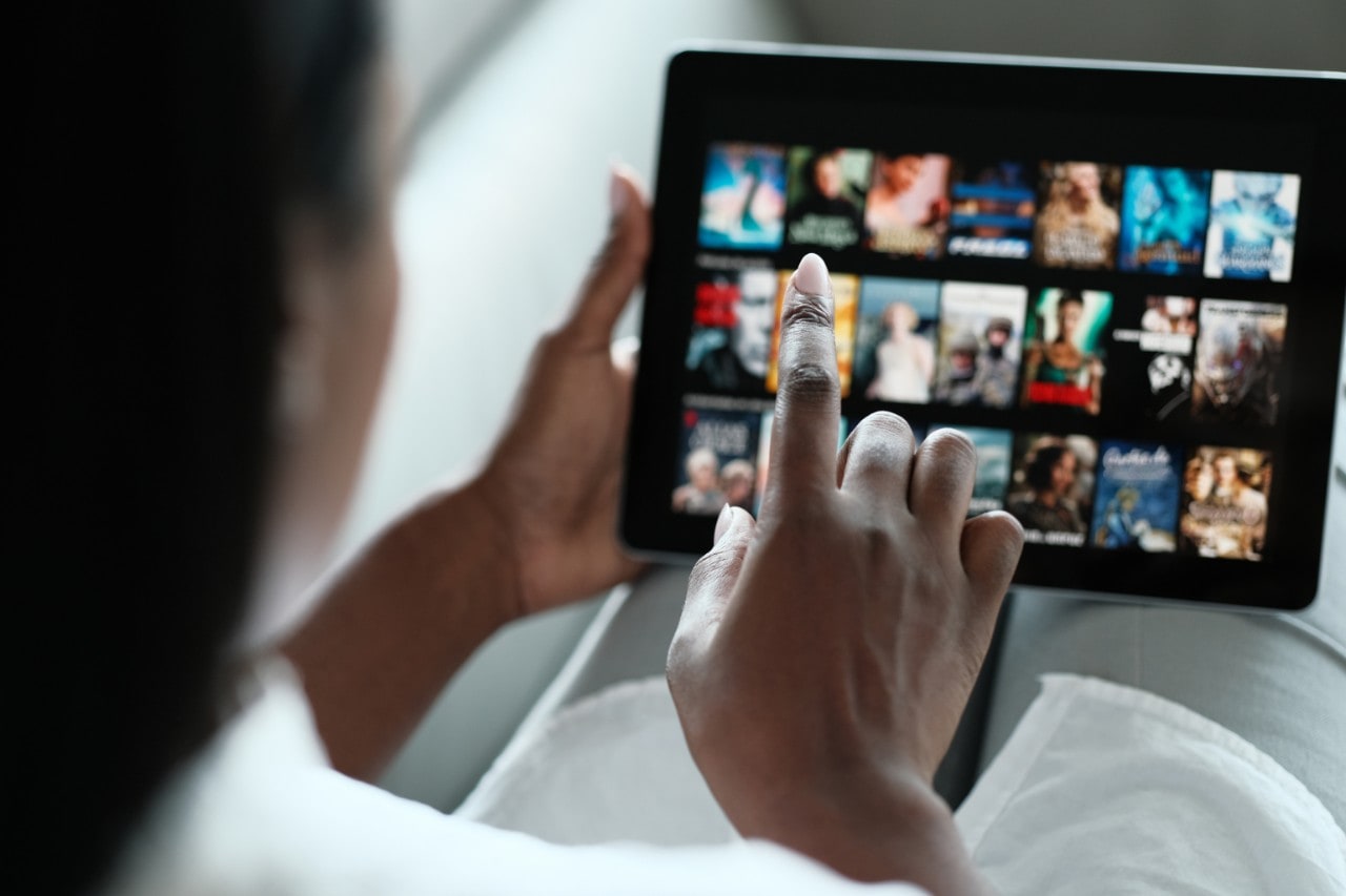 photo of a woman's hand reaching out to tap an iPad to choose a TV show on a streaming platform