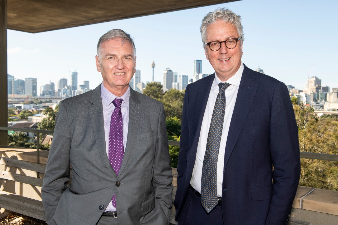 A photo of two men in suits standing in front of a view of the Sydney skyline.
