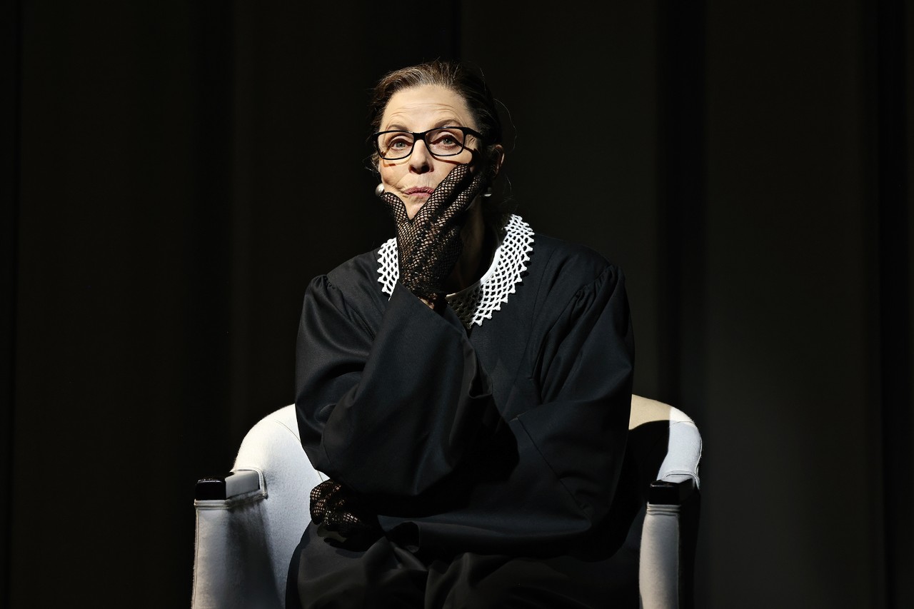 actress Heather Mitchell seated in a chair dressed as America Supreme Court Justice Ruth Bader Ginsburg