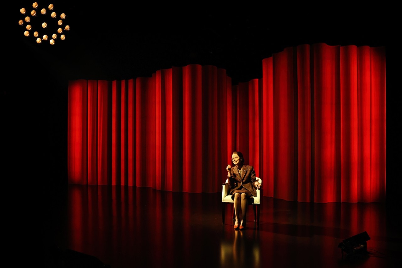 actress Heather Mitchell seated in a chair with a bright red theatre curtain behind her
