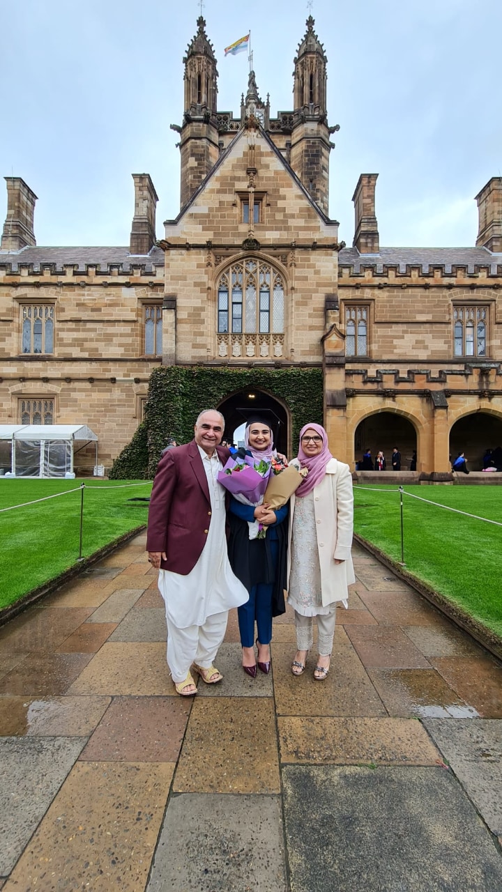 Rameen with her parents side by side at the University of Sydney Quadrangle wearing a black graduation gown and cap.