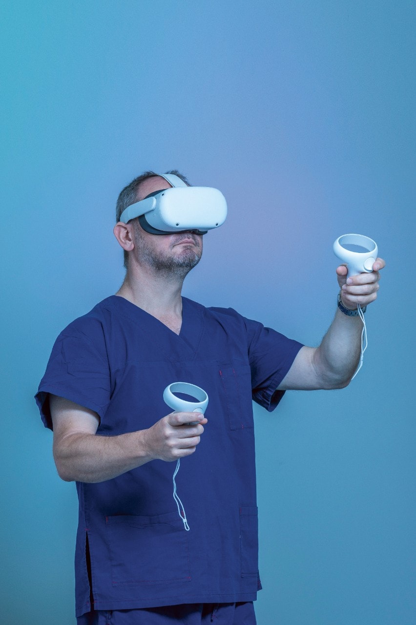 Professor Jonathan Clark in scrubs in front of a blue background with a VR headset on, maneuvering the controls with the handpieces