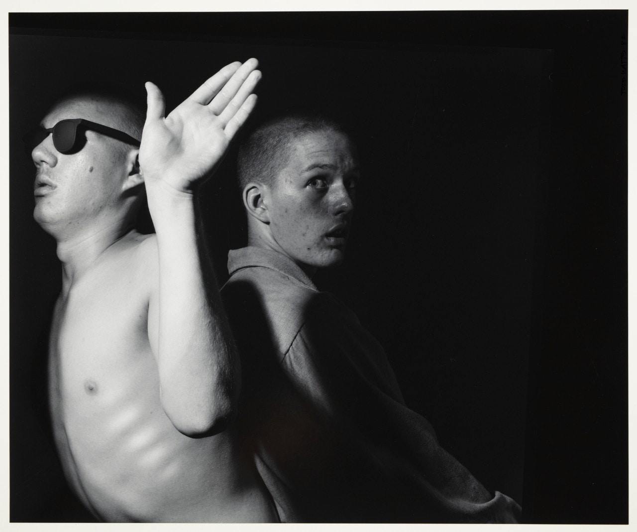 Black and white photo of two men, one on the left is topless, wears sunglasses and has a hand on his shoulderands in the air, the one on the right is obscured and 