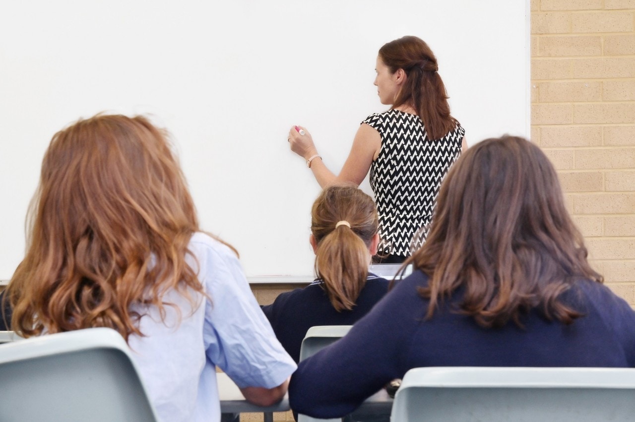 Young students in a classroom face away from the camera to a teacher writing on a whiteboard..