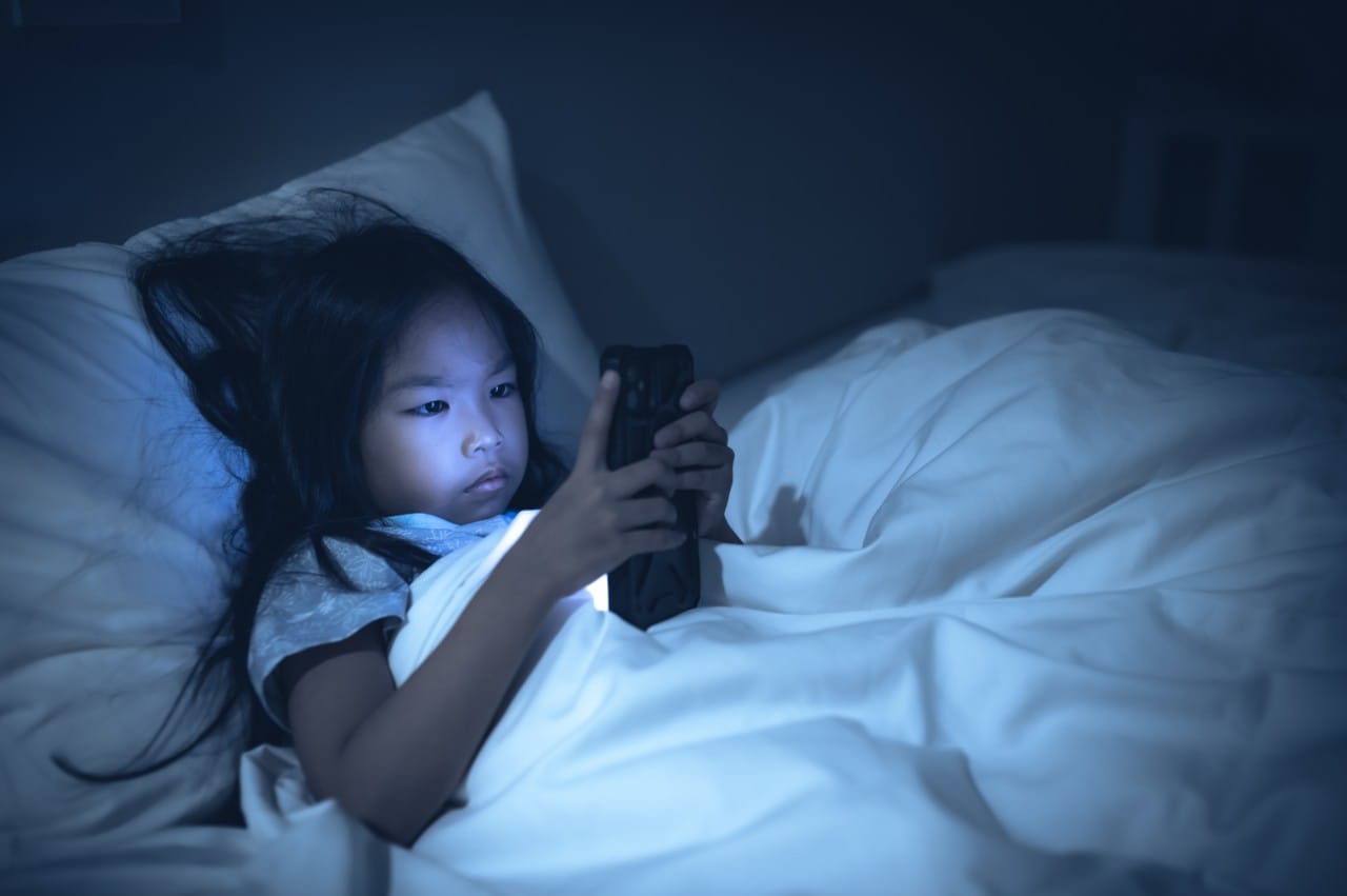 Young girl lies in bed illuminated by the glow of her phone