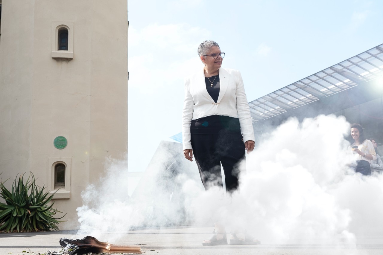 Deborah Cheetham Fraillon is a woman in a white jacket and black pants standing while surrounded by smoke from a smoking ceremony at the Sydney Conservatorium of Music