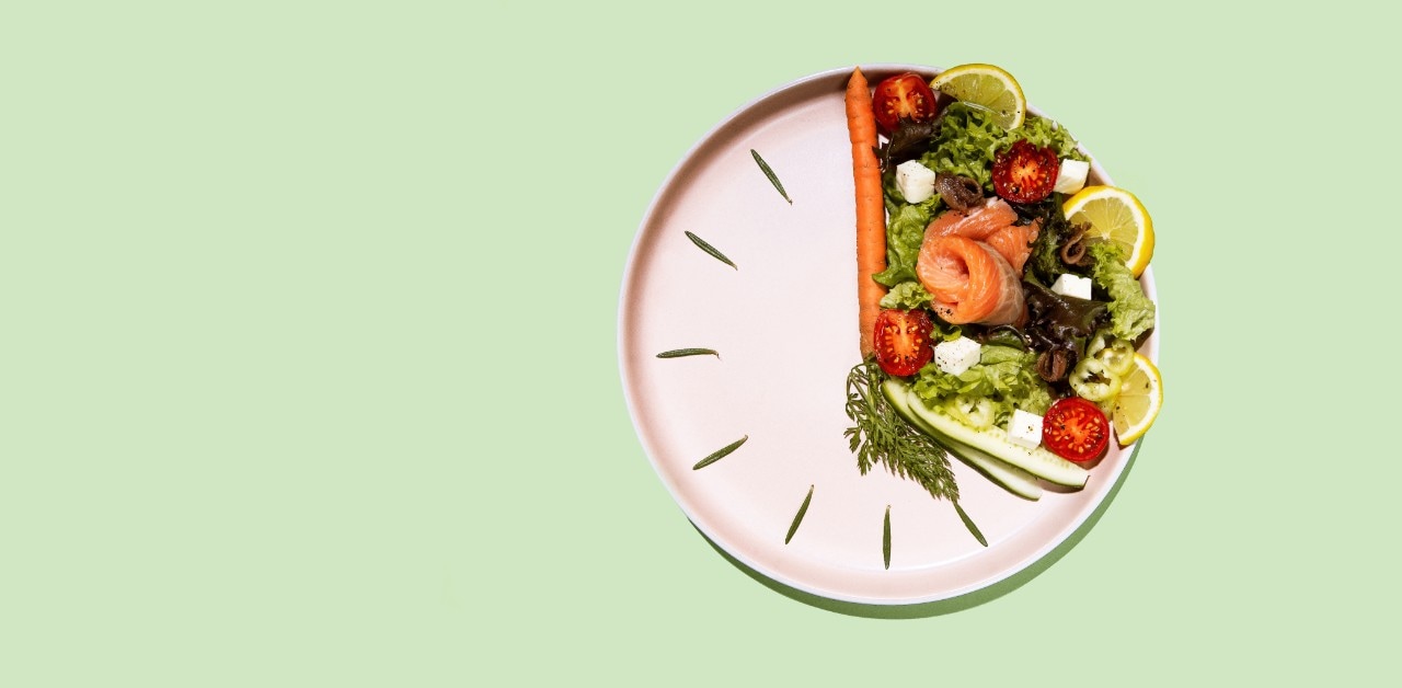 A round plate with a salad sits on a light green background. The salad is arranged in a wedge to take up a third of the plate, borded by carrots and cucumbers to mimic the hands of a clock. Sprigs of rosemary make up the hour markers around the edge of the rest of the plate to signify intermittent fasting and time restrictive diets.