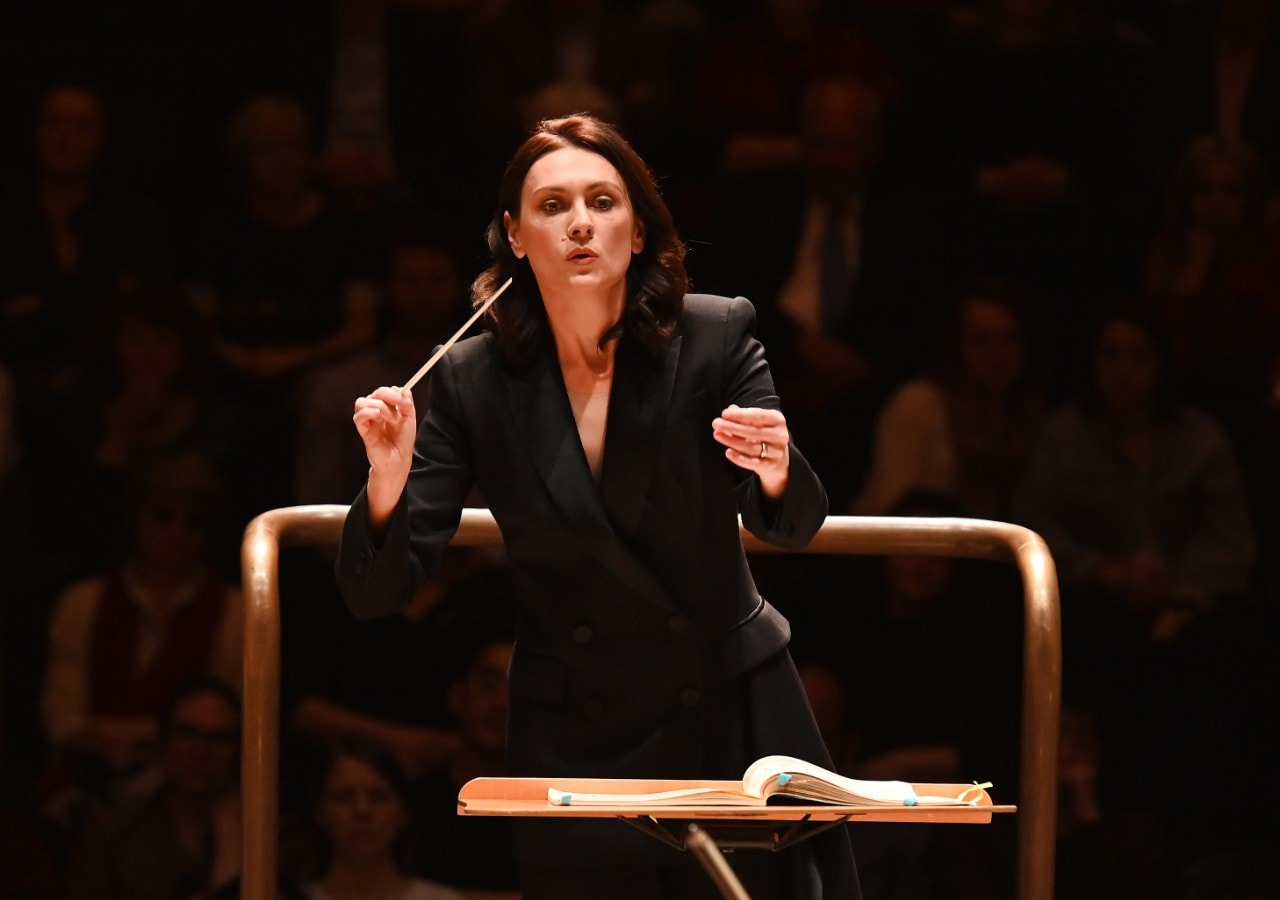 Conductor Natalie Murray Beale on a conducting podium waving her baton, she is looking straight ahead, brown hair, black suit 