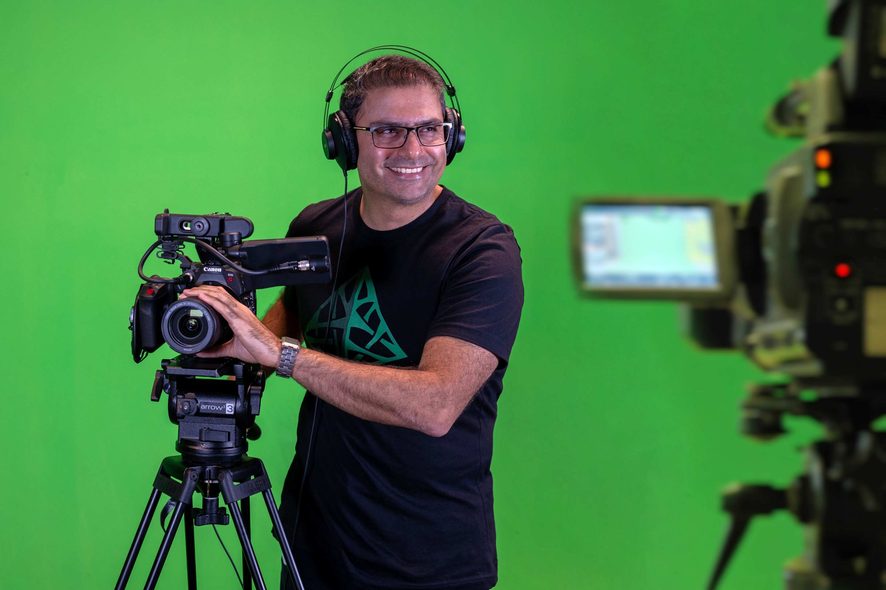 Reza in front of a green screen wearing a headset and holding on to a camera.