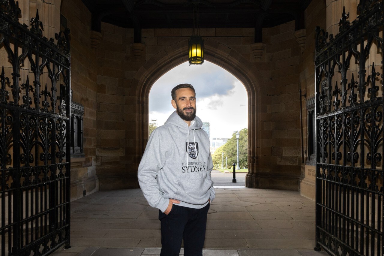 Mathew Demetriou standing the entryway to the Quad in a grey University of Sydney hoodie.