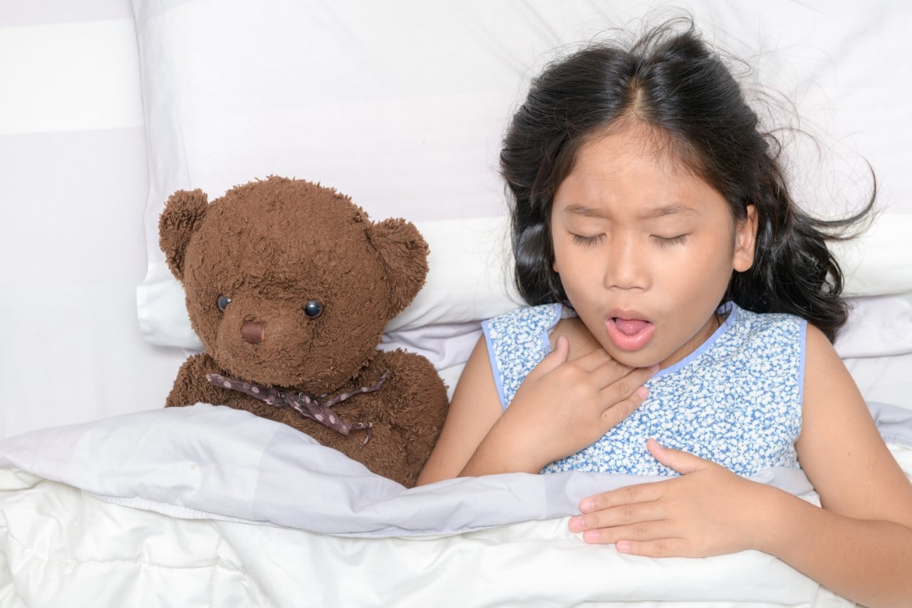 A young girl coughs while lying sick in bed with her teddy bear.