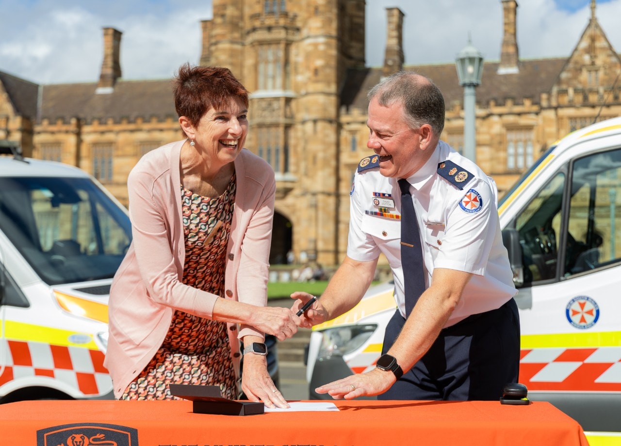 Robyn Ward and David Dutton sign an agreement in front of two ambulances with the University Quad in the background.