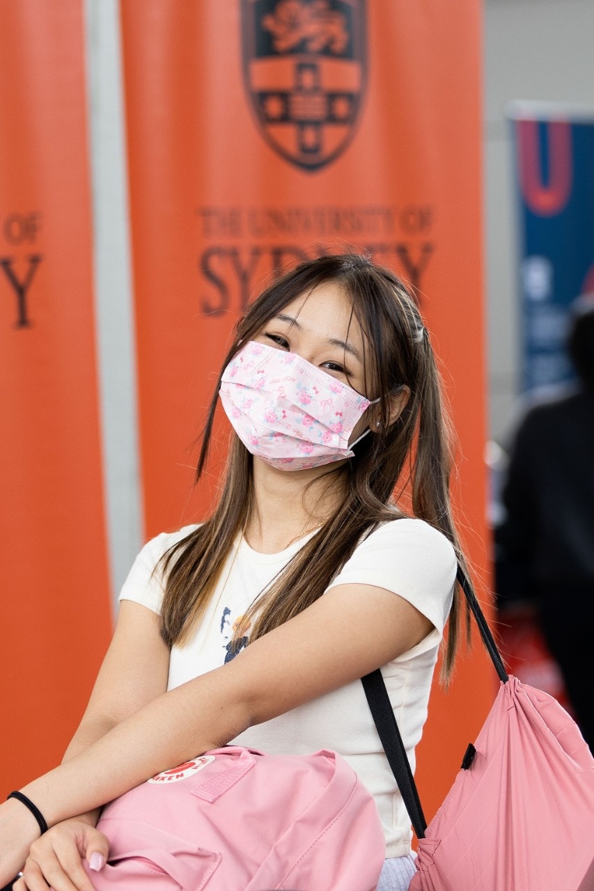 A girl with a pink mask and pink luggages smiling in front of the USYD banner at the airport