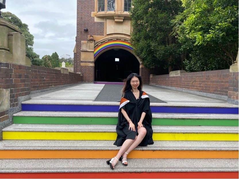 Yuning sitting on rainbow stairs outside Manning wearing graduation robes