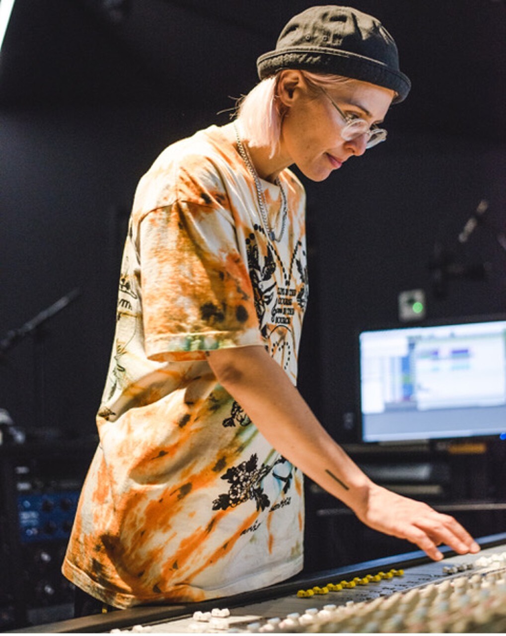 Jane Arnison photographed in a music studio using a sound mixing machine. She wears a tie-dye tshirt, a black bucket hat and clear-rimmed glasses.
