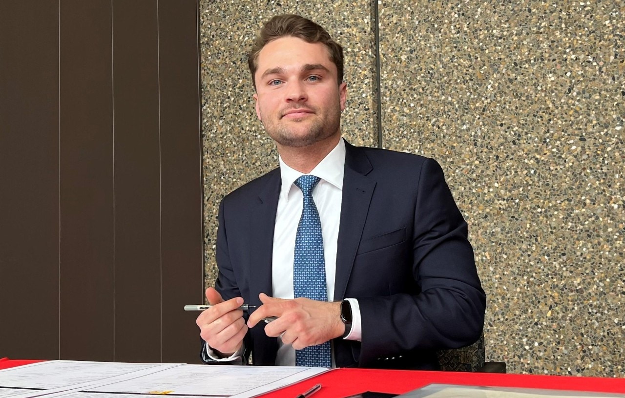 Man in a suit and tie seated behind a table, holding a pen and about to sign a document.