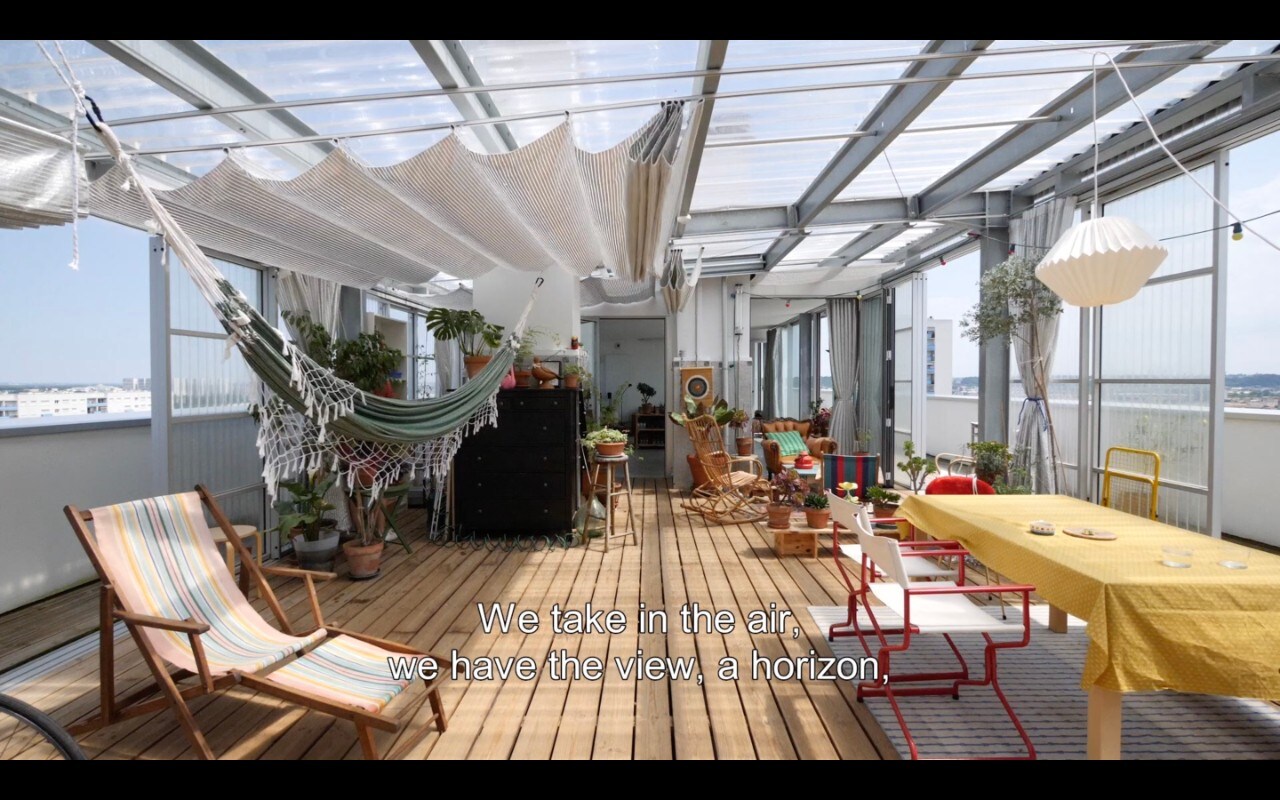 A still from a video showing a rooftop apartment. Subtitles read: "We take in the air, we have the view, a horizon,"
