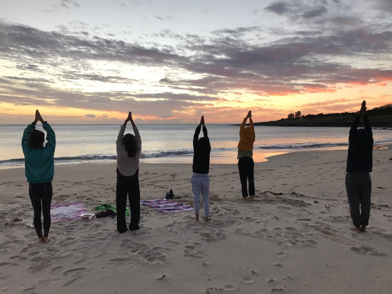 Five people standing on a beach doing making a prayer guesture with their arms up facing the ocean.
